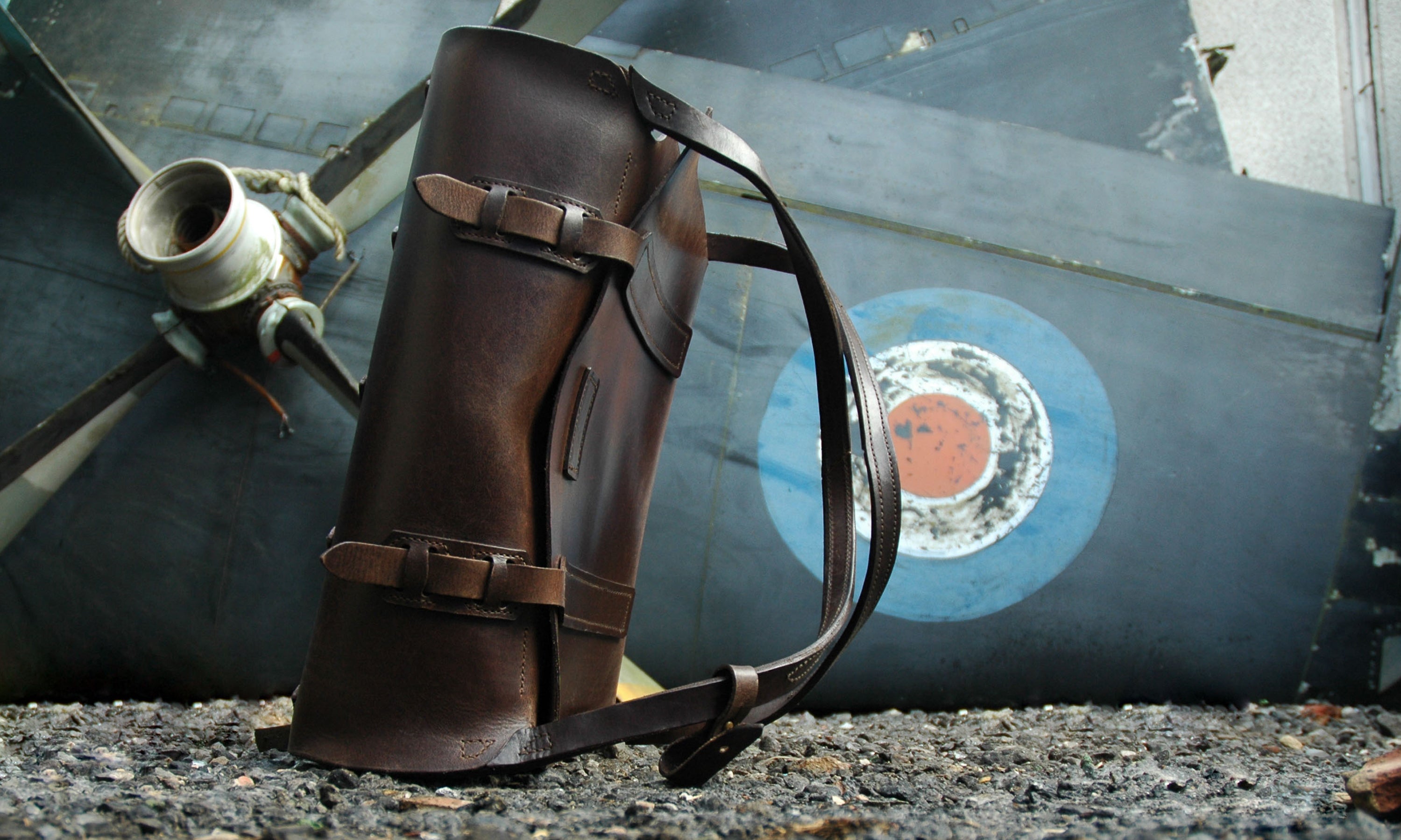 Vintage style backpack for overnight and weekend travel. The backpack sits in an scrapyard with old RAF planes.
The 'Parachuter Bag' is handmade by Garvan de Bruir and features classic leather details in veg-tanned cow hide.
