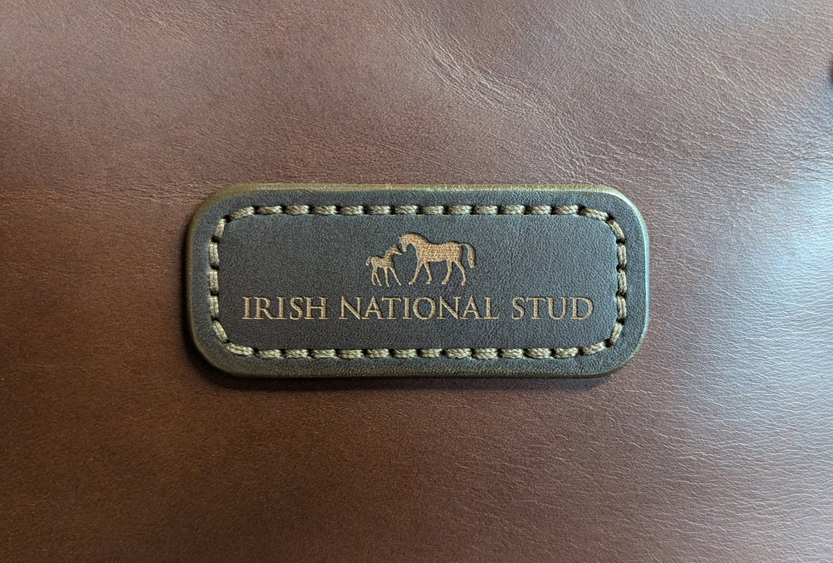 Rectangular full-grain leather brand tag with 'Irish National Stud' branding on a brown leather bag