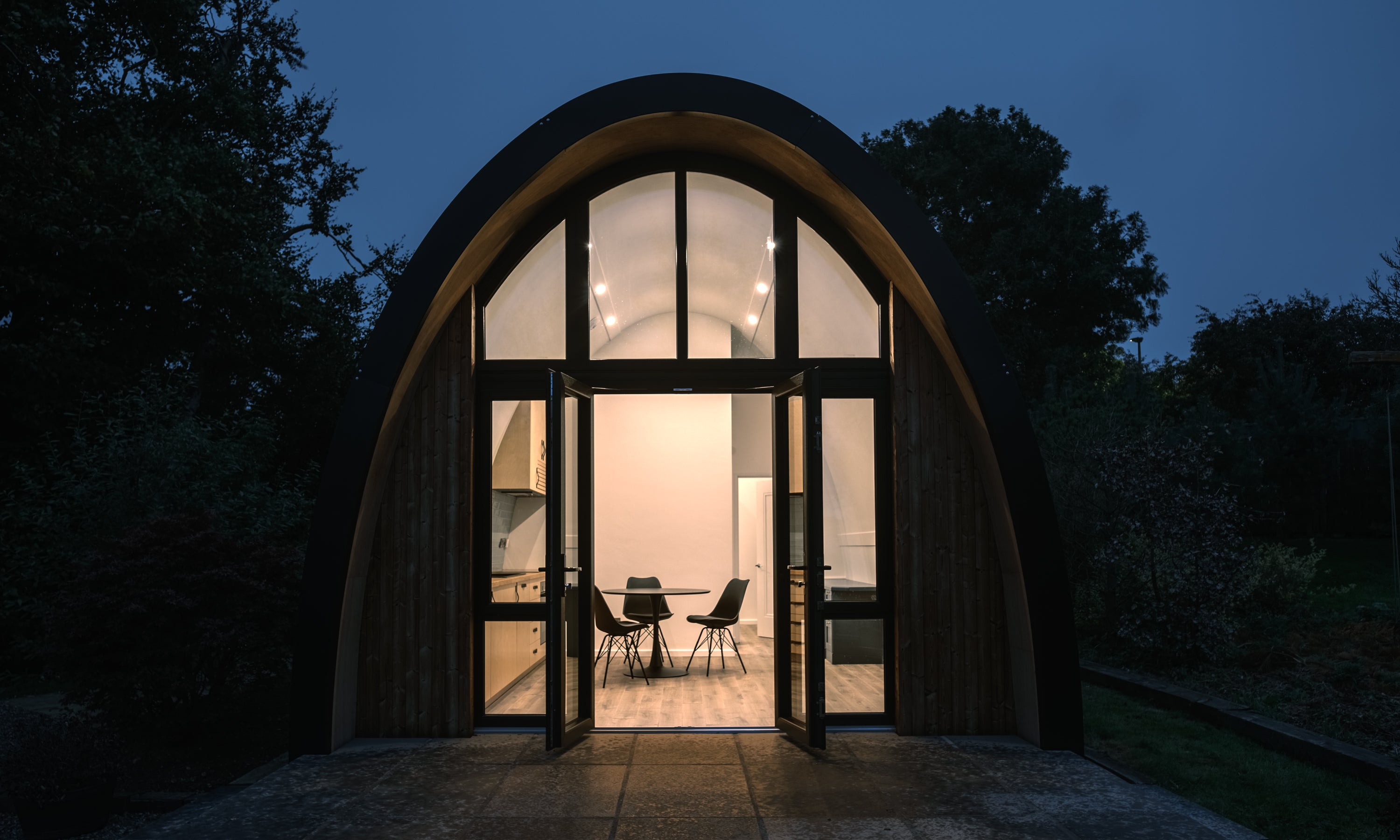 A small modular dwelling with a curved prefabricated roof design. The front of the building has full height glazing with a timber surround. 